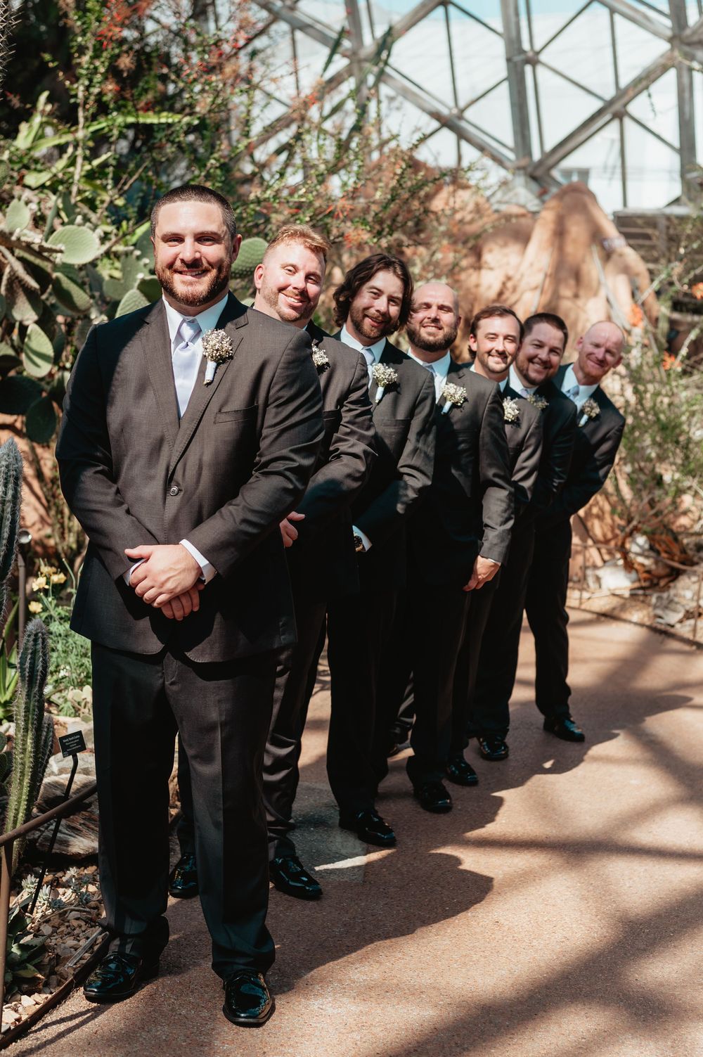 Groomsmen in dark suits with boutonnieres lined up in a greenhouse botanical garden setting for a wedding photo.