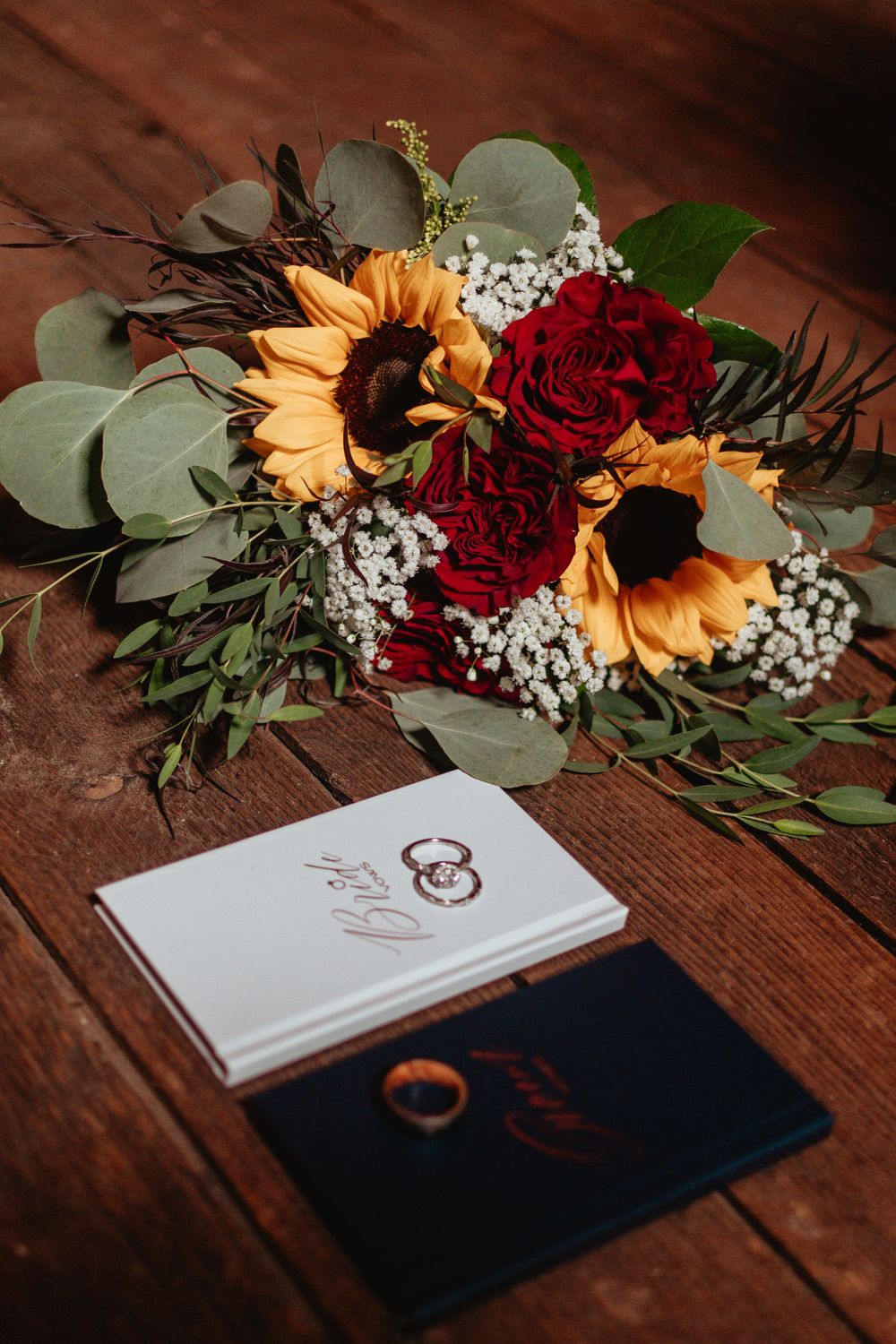 Wedding stationery with gold lettering alongside a fall bouquet of sunflowers, red roses and eucalyptus on wooden table.
