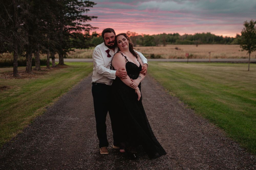A couple stands on a gravel path at sunset, with trees and fields in the background.