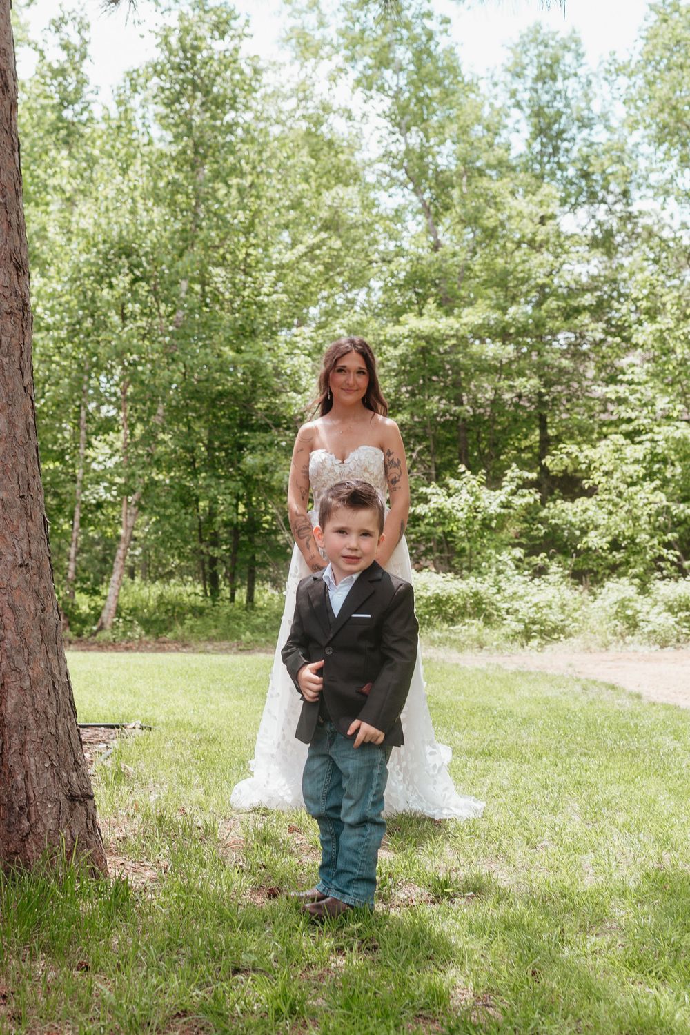 A bride in a white wedding dress stands behind a young boy wearing a dark suit jacket on grassy woodland grounds.
