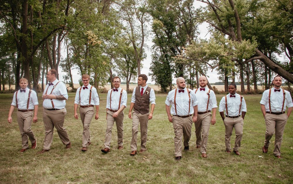 Groomsmen in suspenders and khakis stand in a line on grassy lawn for casual outdoor wedding photo.