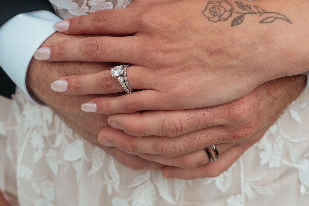 Close-up of two hands with wedding rings and a small tattoo resting on white lace fabric.