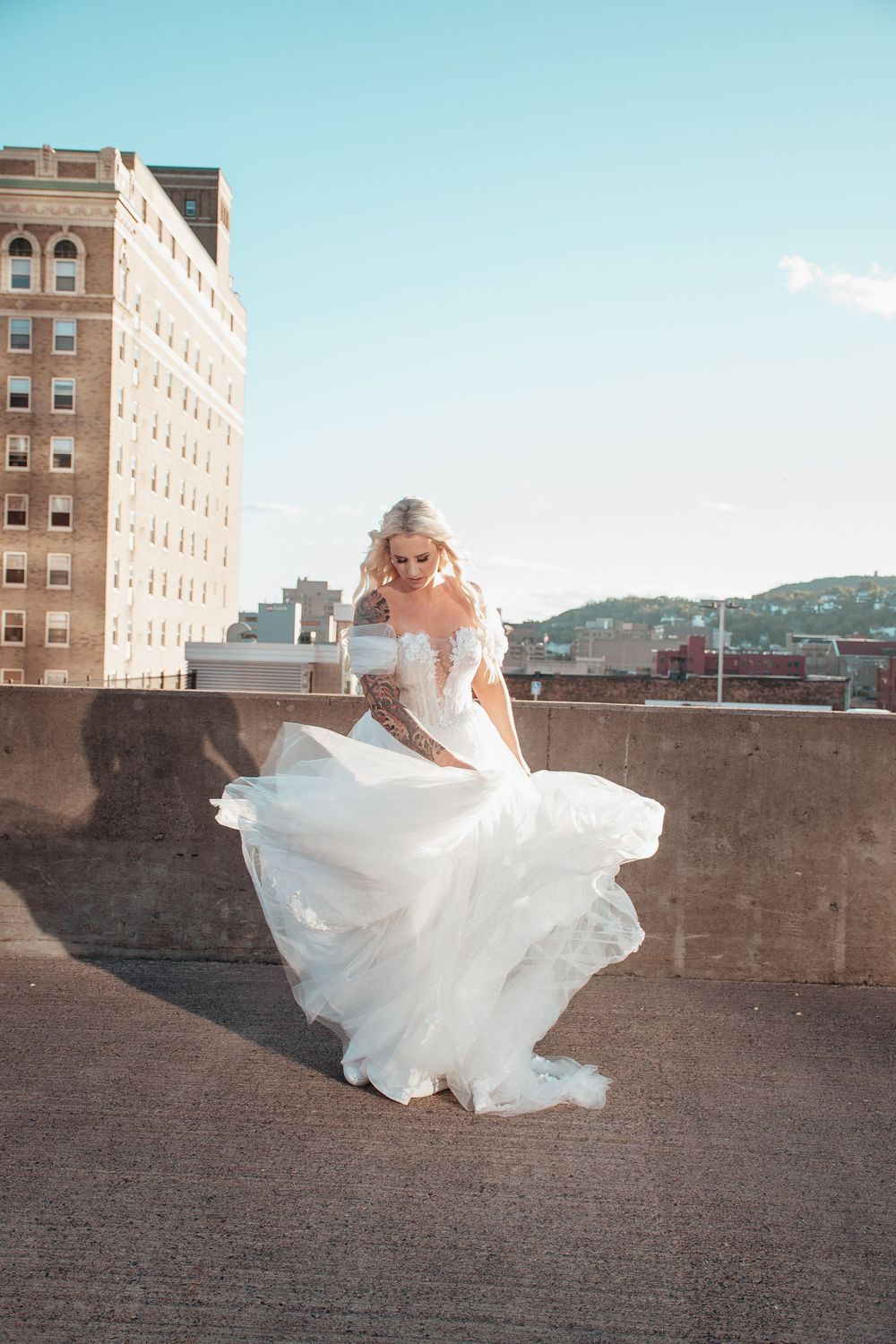 A bride in a flowing white gown twirls on a rooftop with city buildings in the background.