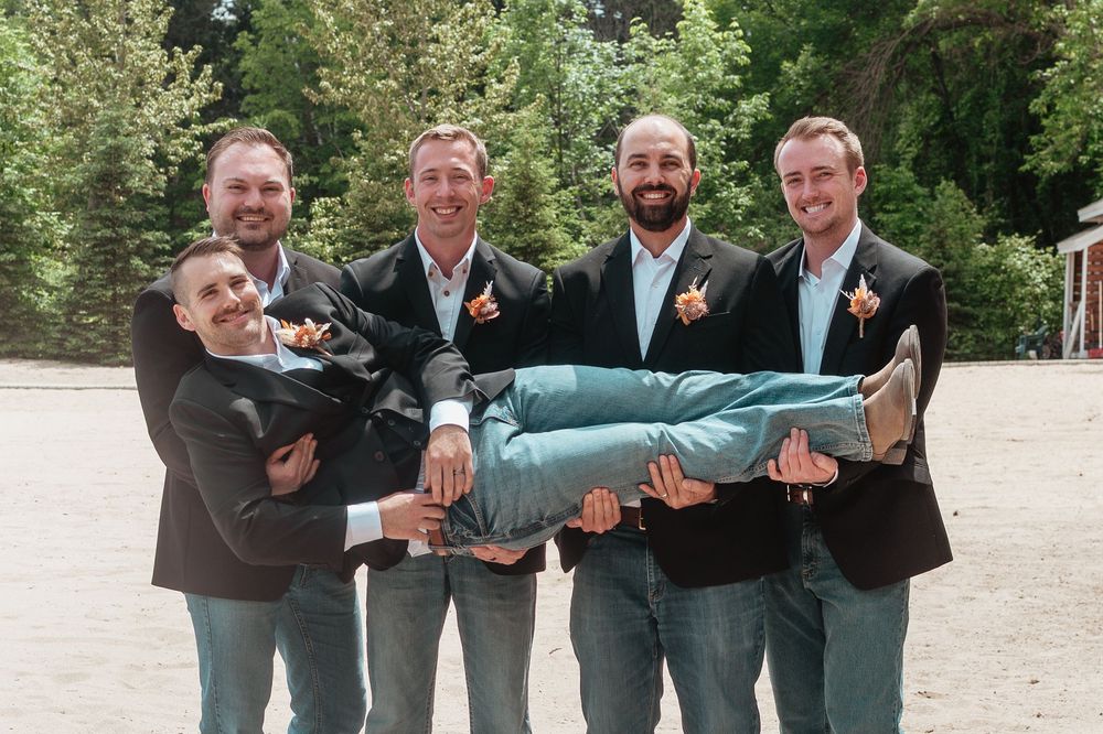Four groomsmen in tuxedos playfully lift each other in matching poses outdoors during a wedding photo session.