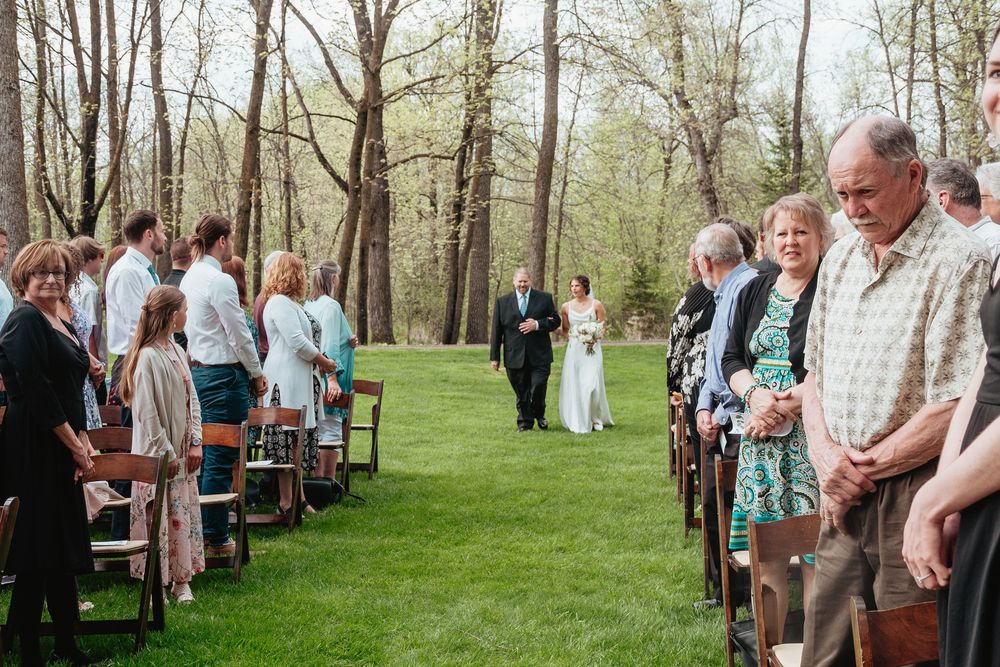 Wedding guests gather outdoors on a grassy lawn surrounded by trees during a spring celebration event.