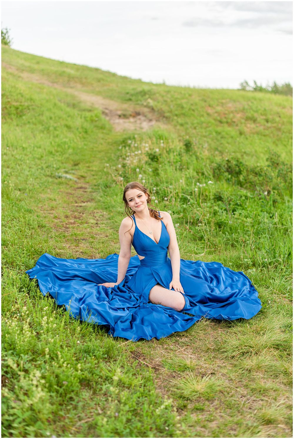 Elegant blue gown spreads across grassy trail in outdoor portrait session.
