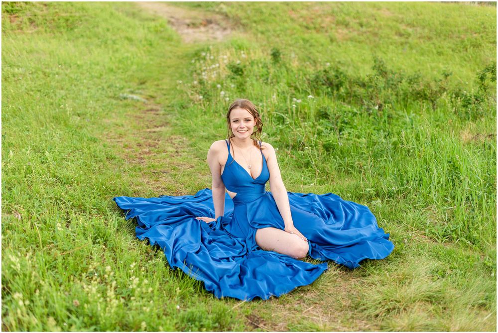 A blue evening dress spreads elegantly across green grass in an outdoor portrait setting.
