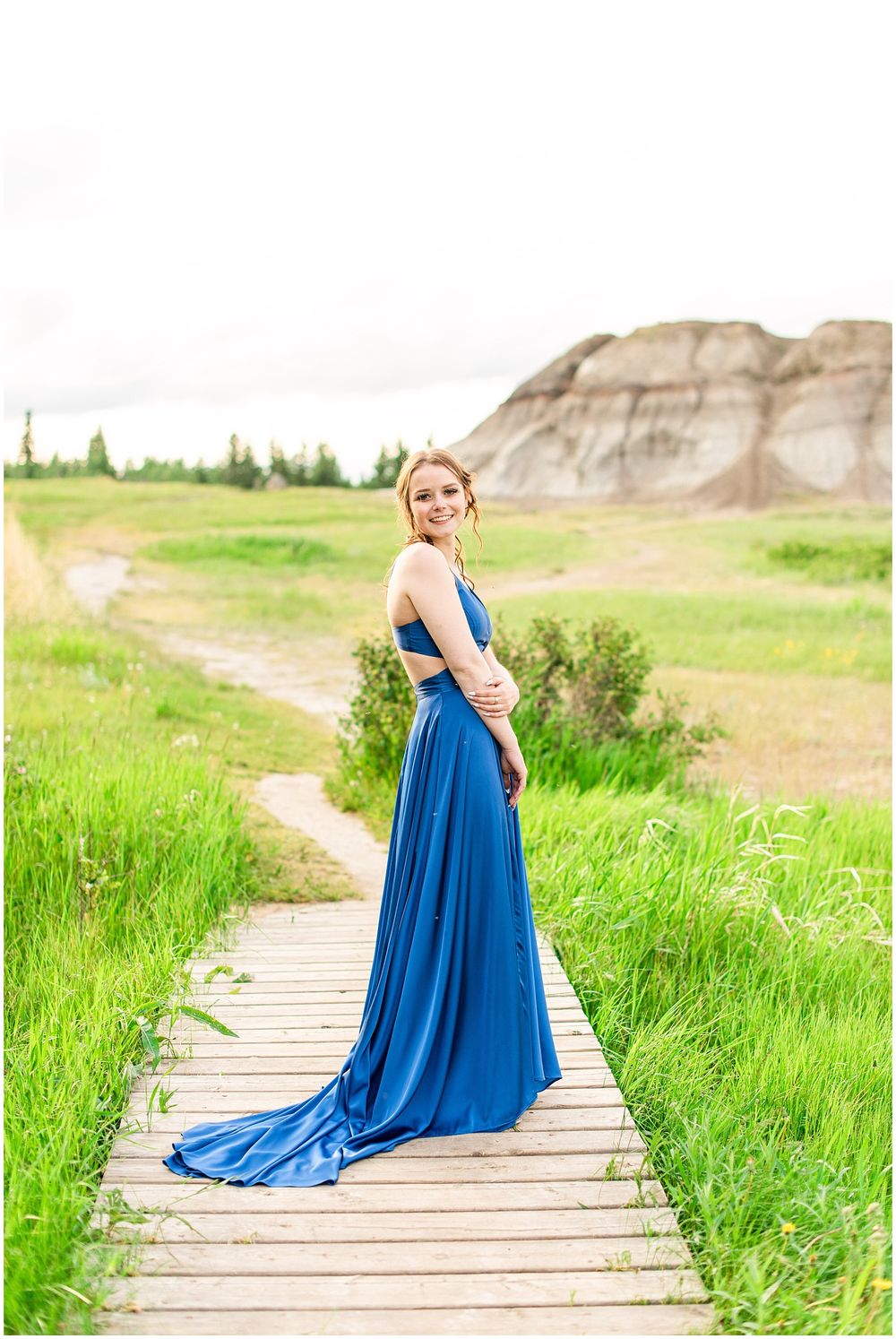 Striking blue formal dress with train photographed on wooden boardwalk through prairie landscape.