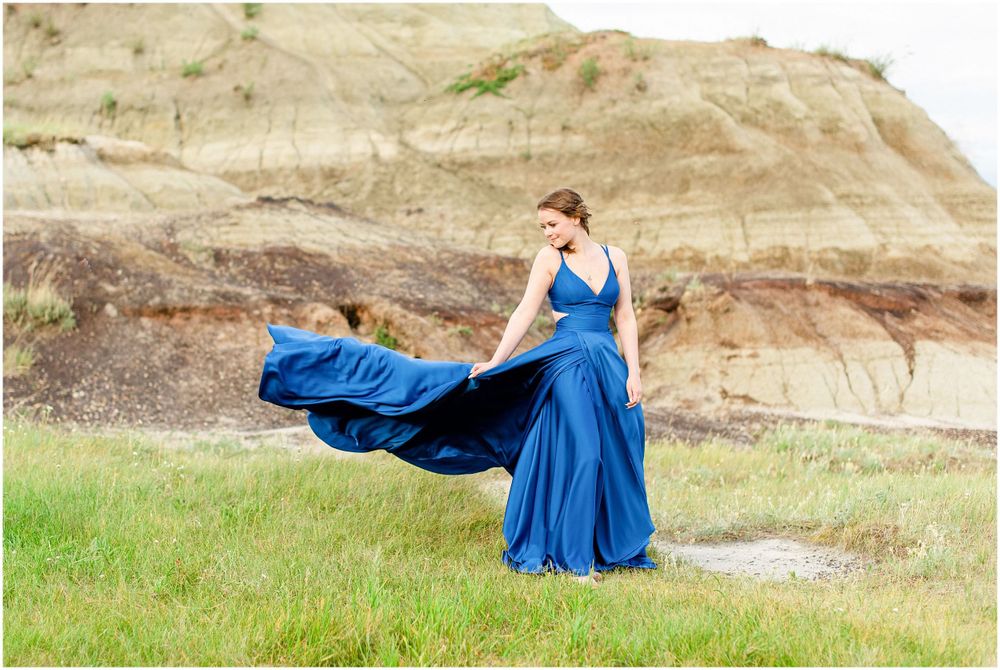 Elegant flowing royal blue formal dress billowing in the wind against rocky hillside backdrop.