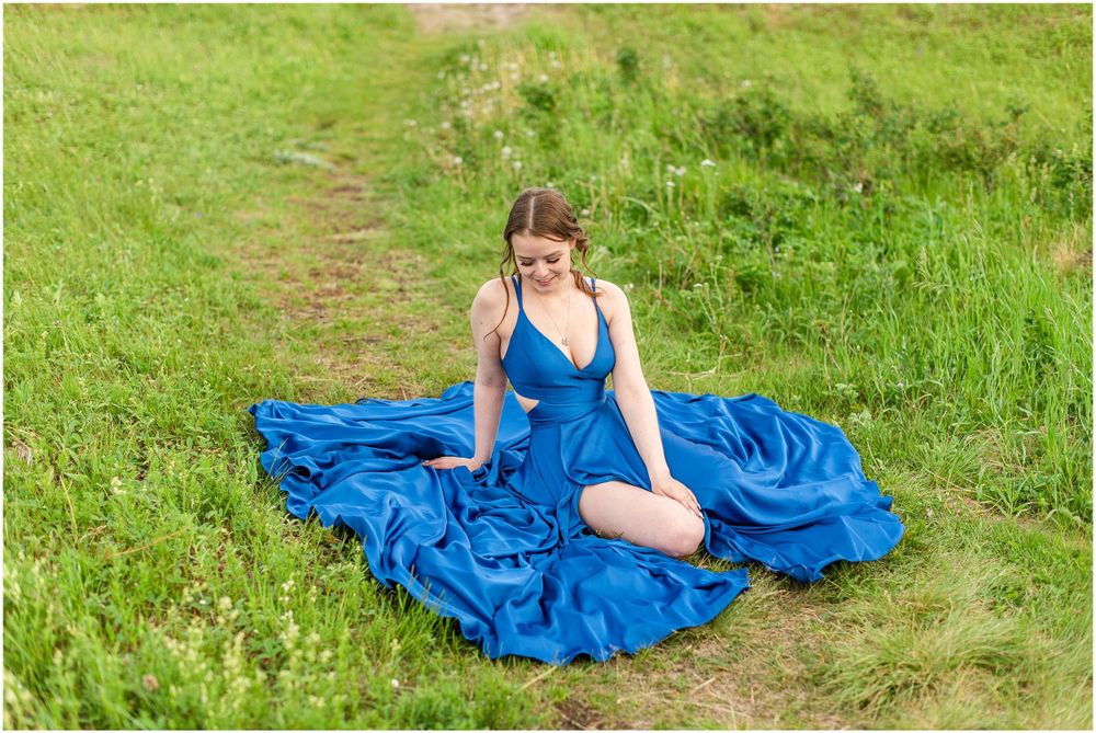 Elegant blue gown with flowing fabric spreads across grass in outdoor portrait setting.
