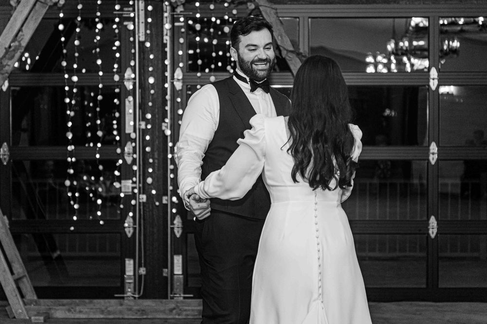 A black and white photo of a couple embracing during their wedding celebration with twinkling lights in the background.