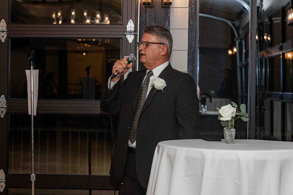 A speaker in a black suit and tie holds a microphone while giving a speech at an elegant indoor reception venue.