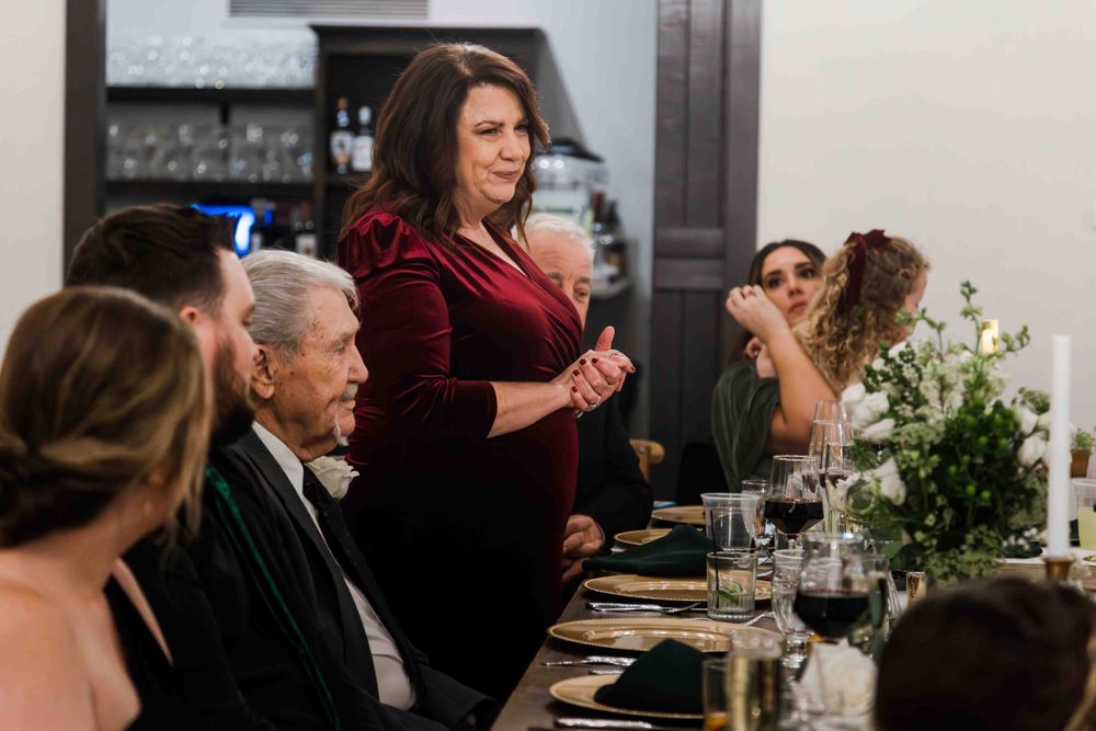 People gather at an elegant dinner table with candlelight and floral centerpieces while someone stands to give a speech.