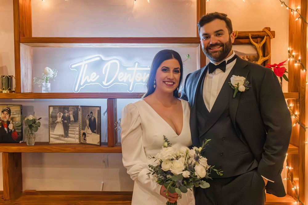 Newlyweds pose in front of an illuminated neon sign at their reception with elegant white bouquet and formal attire.