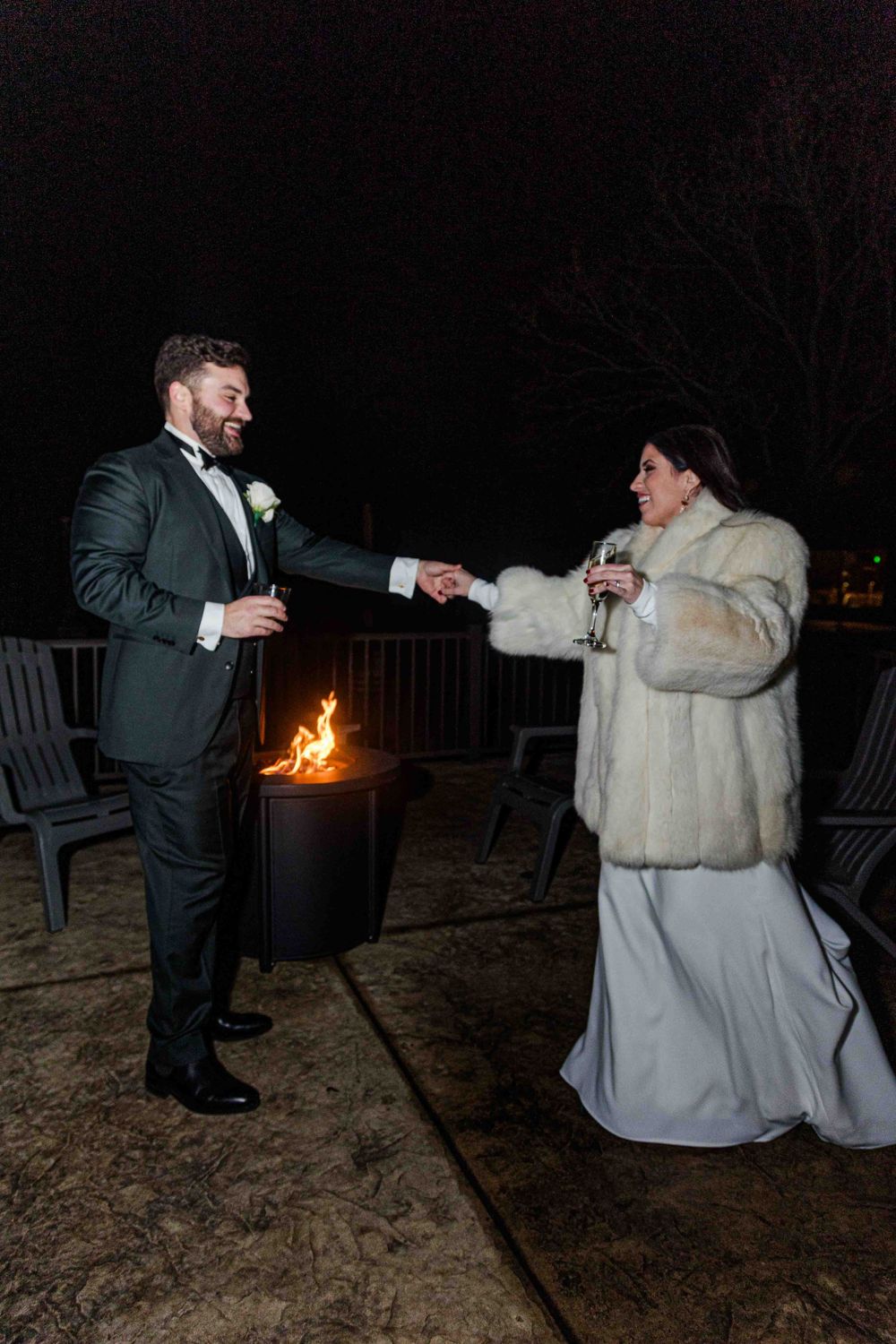 A bride in white dress and fur coat dances with groom in tuxedo near outdoor fire pit on winter evening.