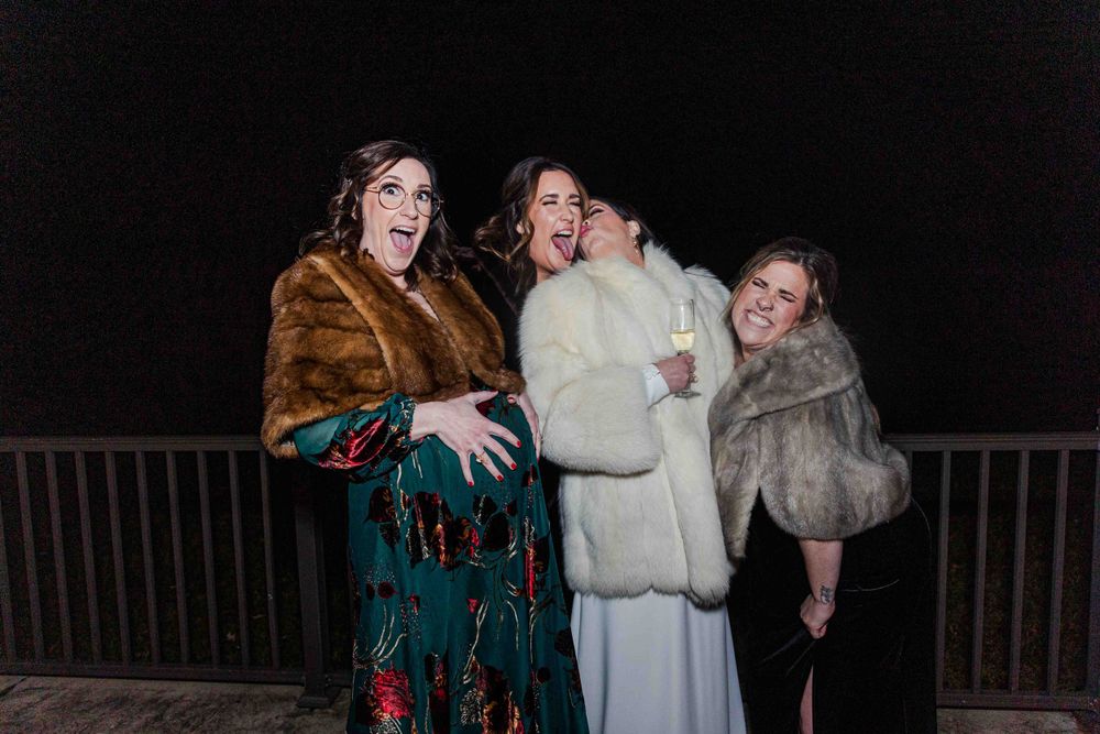 Three people in fur coats laughing together at night on a balcony railing.