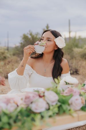 Woman in white dress enjoying tea at outdoor desert setting with pink floral arrangements.