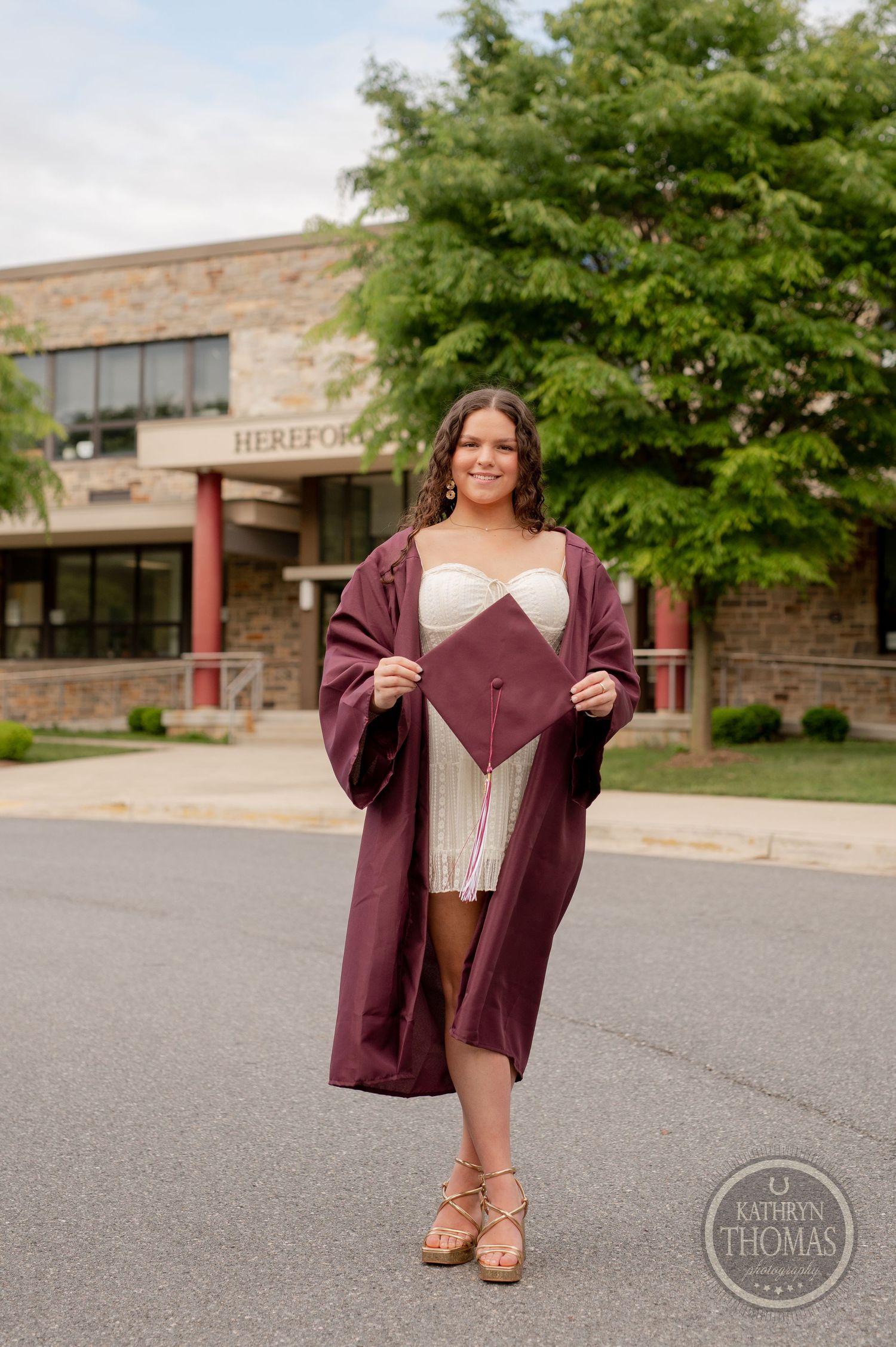 Graduate poses in burgundy cap and gown with white dress outside stone school building.