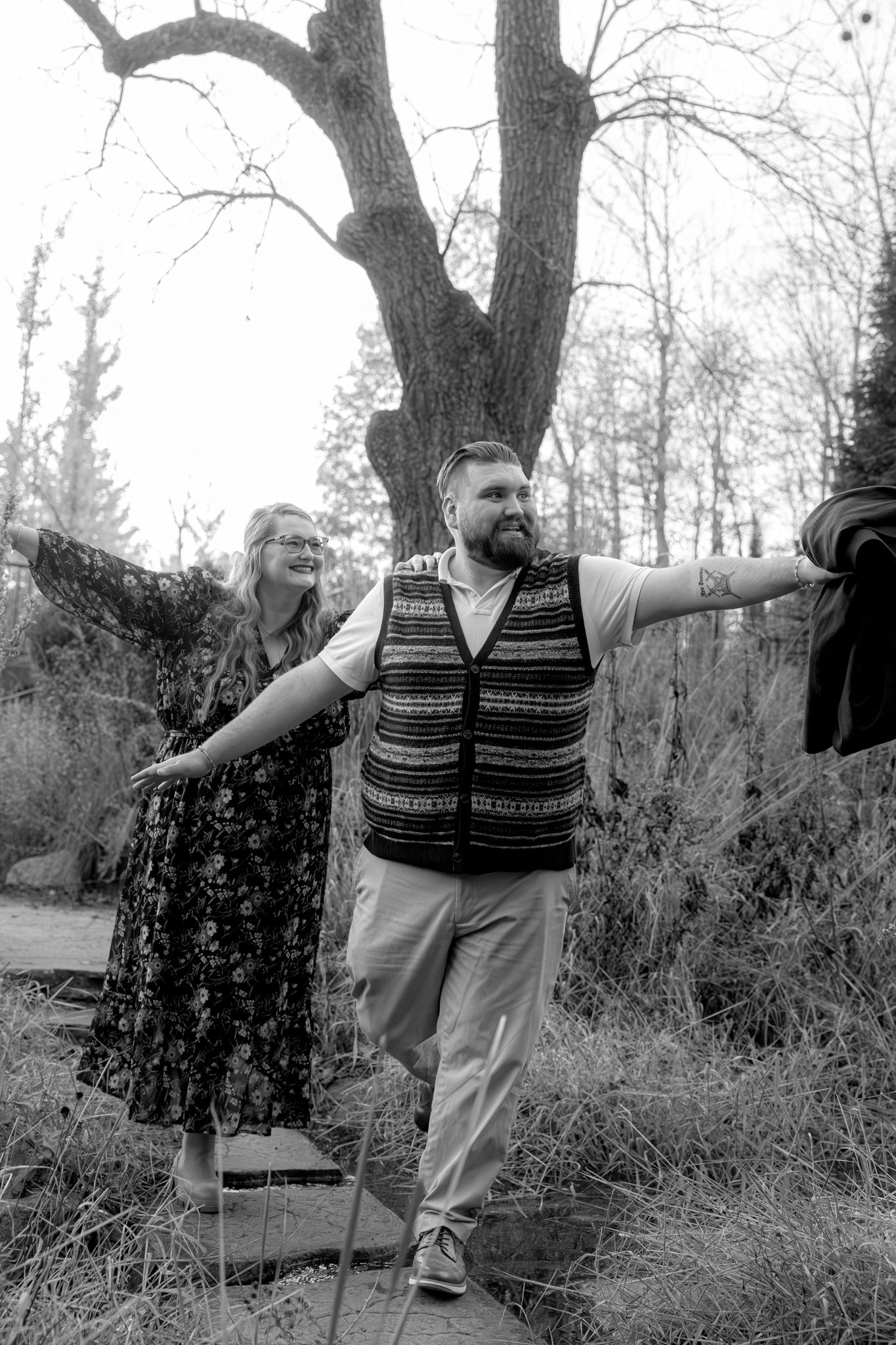 Black and white photo of couple posing playfully outdoors near large tree.