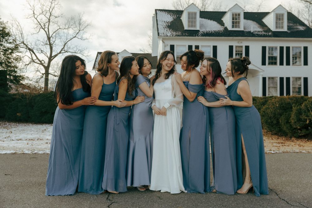 Bridesmaids in flowing dusty blue dresses stand together with bride in white gown outside stately colonial house.