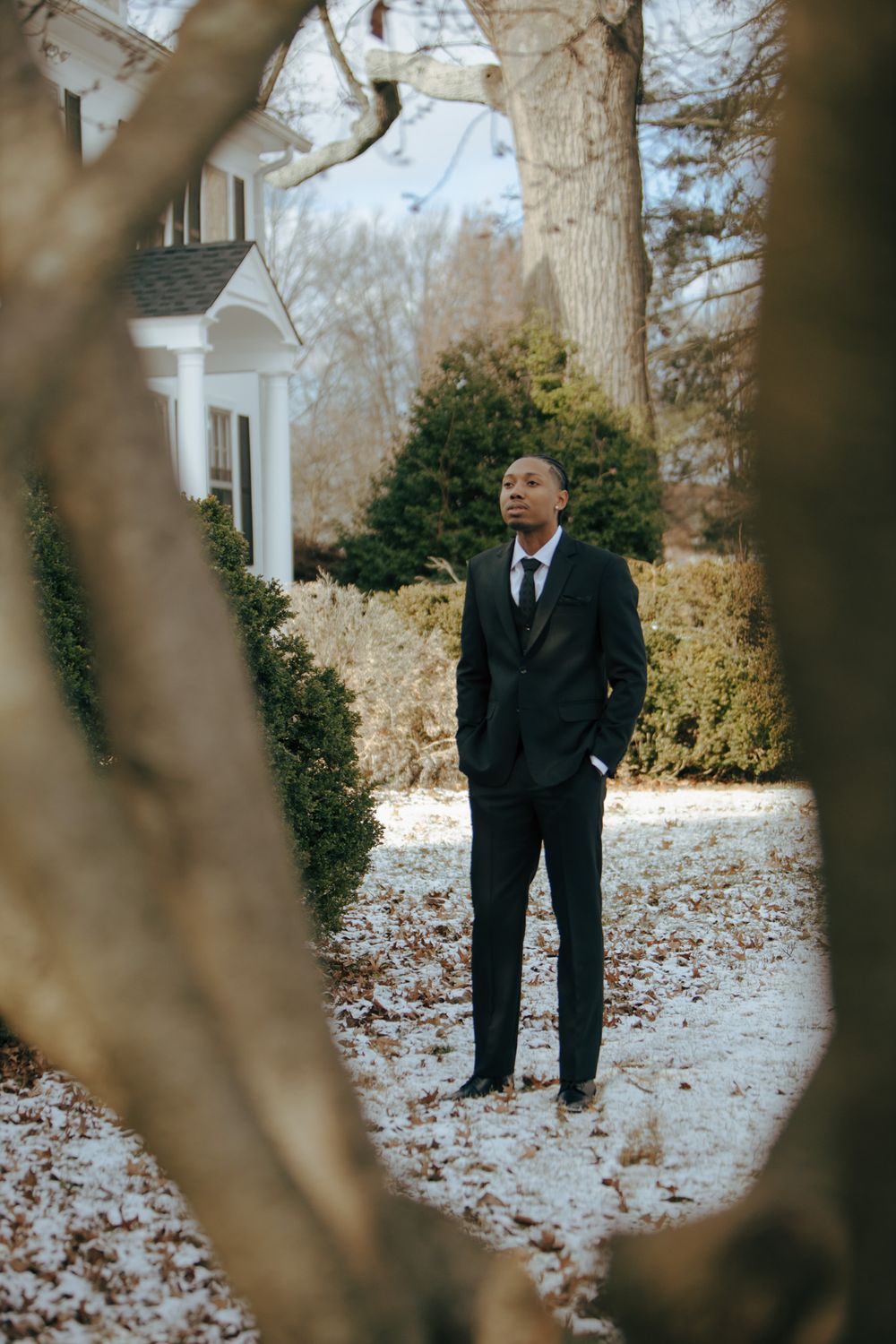 A person in a formal black suit stands near a house on a winter day with snow-covered ground and trees.