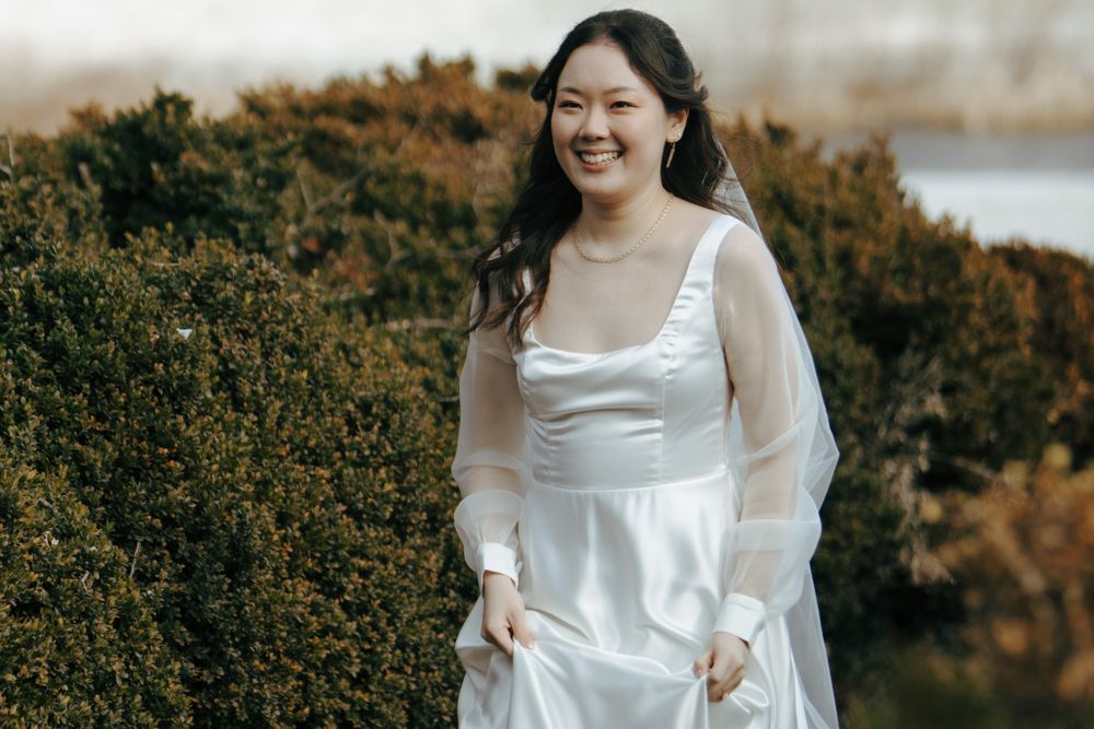 A person in a flowing white dress with sheer sleeves poses outdoors against lush green foliage on an autumn day.