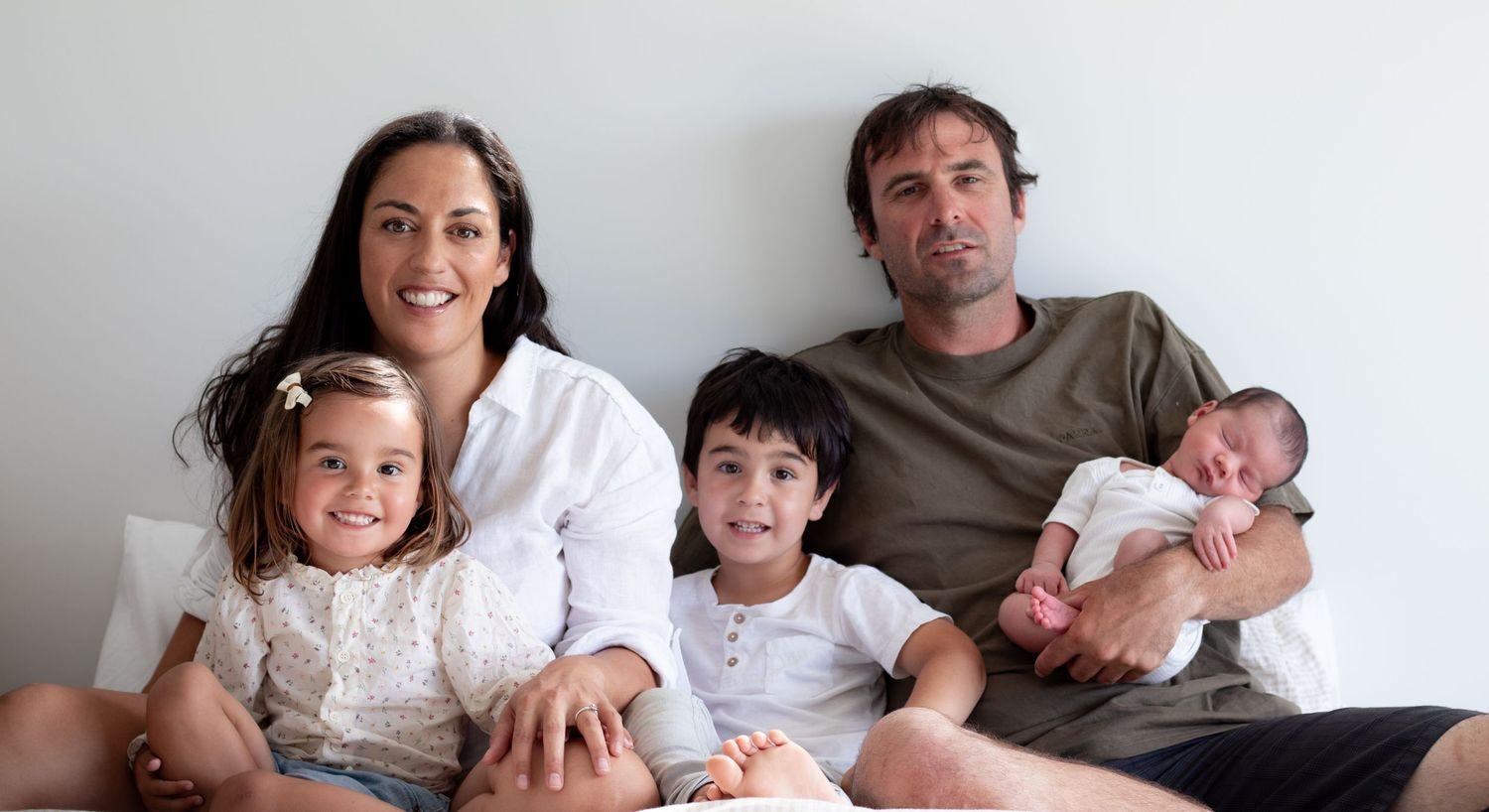 A family of five sits together on a bed nagainst a light colored wall.