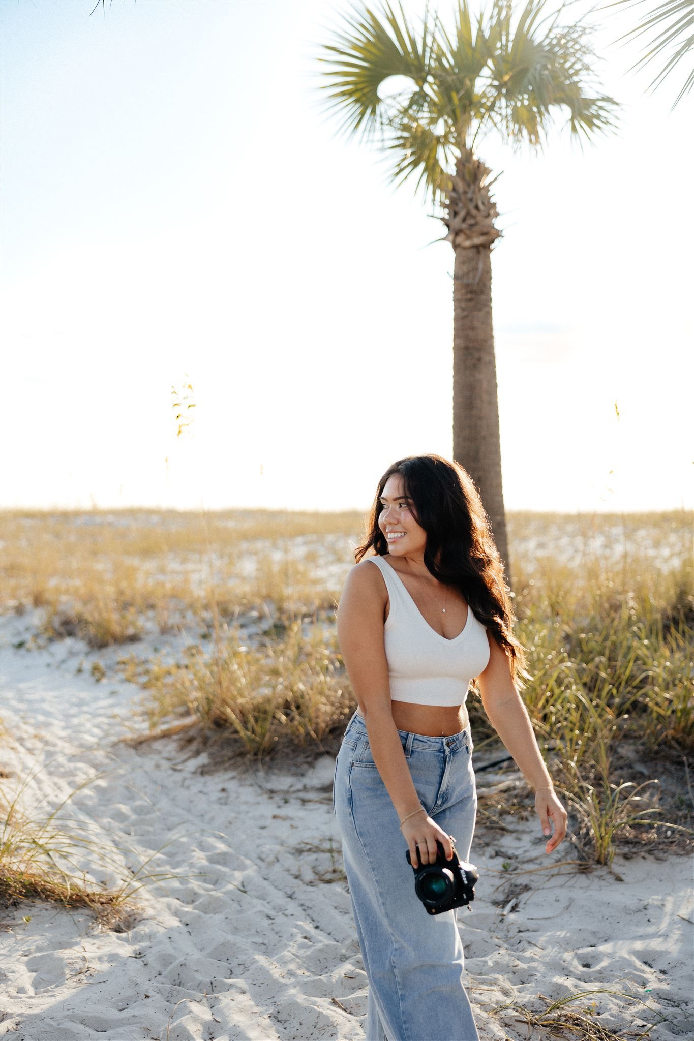 Woman in casual attire holding camera by palm tree on beach path.