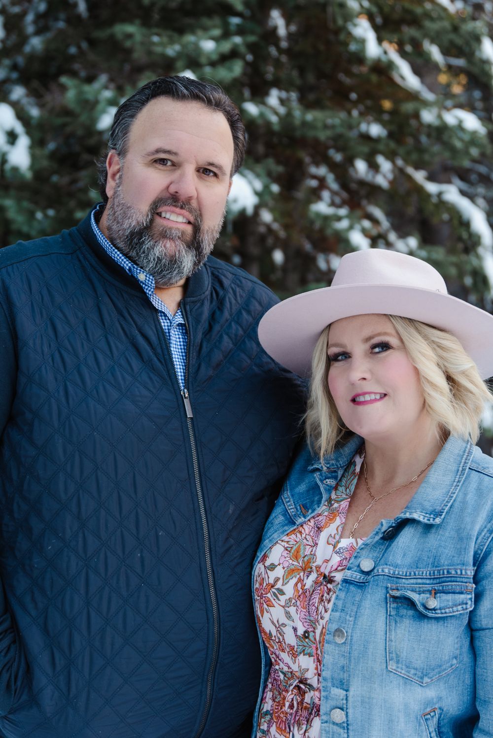 A couple poses for a winter portrait against snowy evergreen trees.