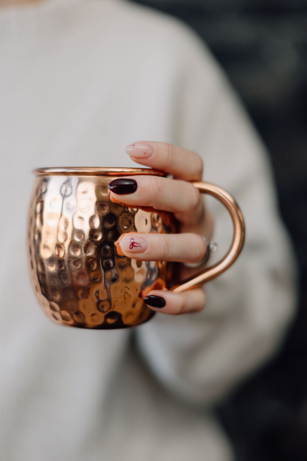 A hand with black and gold manicured nails holds a hammered copper Moscow mule mug against a blurred white sweater.