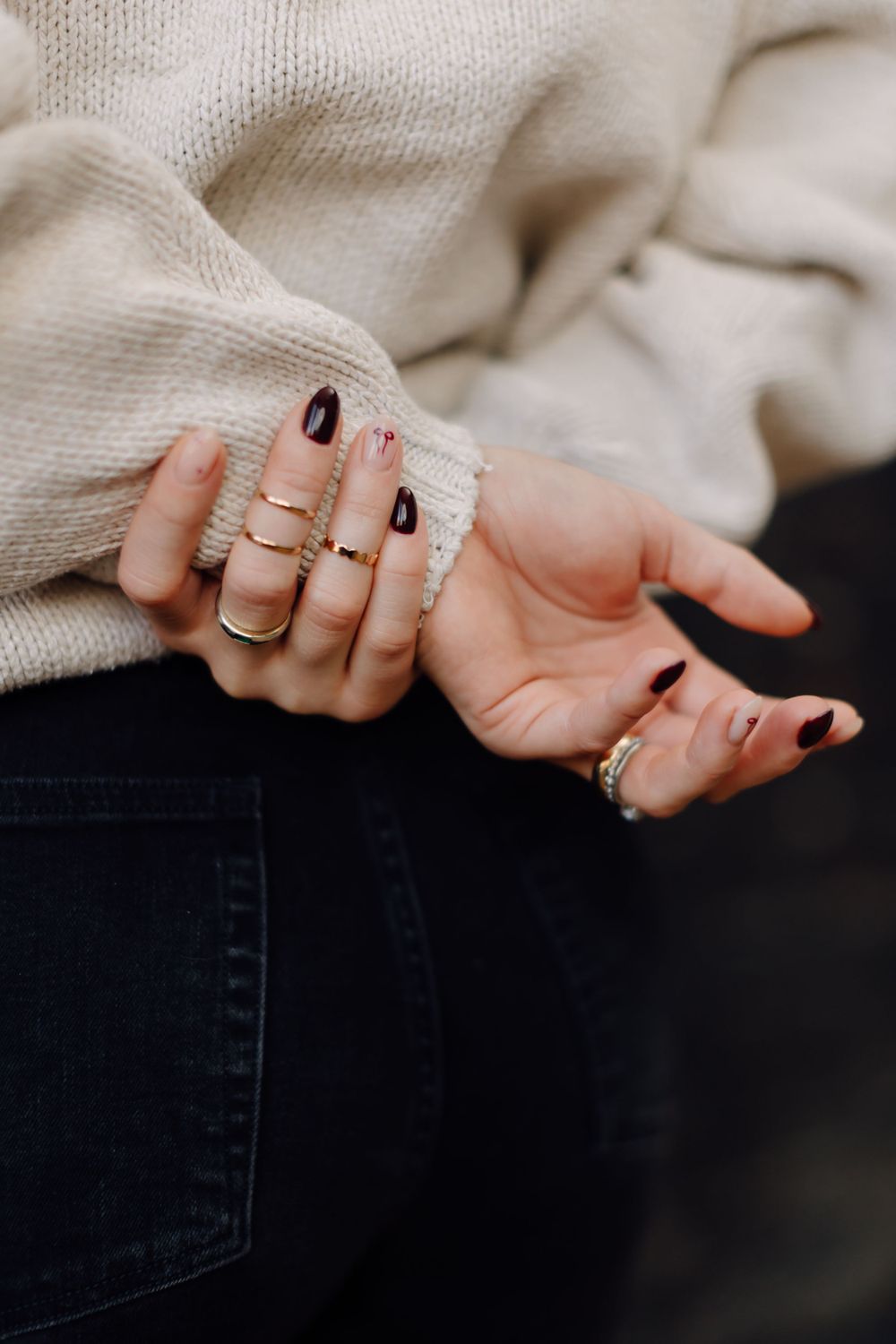 Close-up of hands wearing black nail polish and multiple delicate gold rings against a cream sweater and dark jeans.