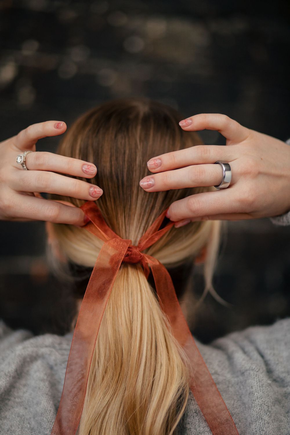 Hands wearing silver rings tie a sheer orange ribbon around a blonde ponytail against a dark background.