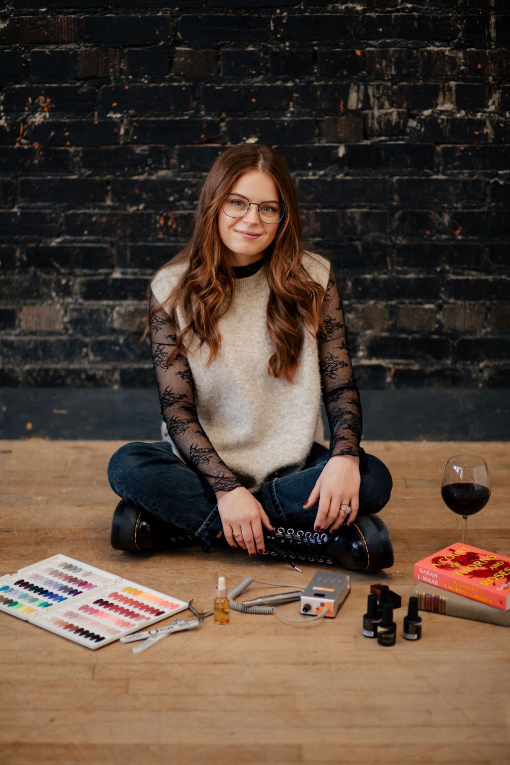Artist sits cross-legged on wooden floor with art supplies, paint swatches, and glass of wine against dark brick wall.