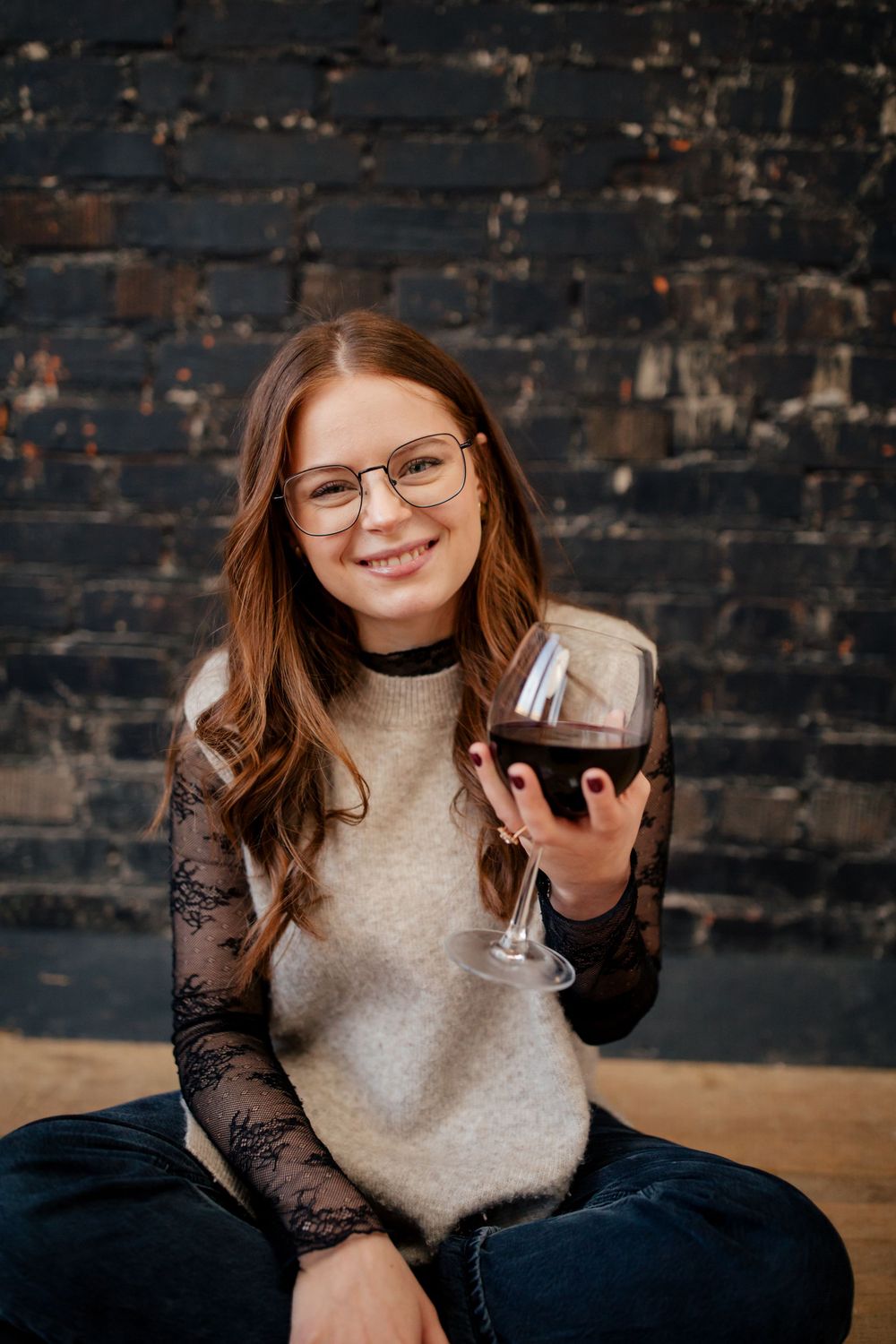 A person in glasses and a beige sweater with lace sleeves holds a glass of red wine against a dark brick wall.