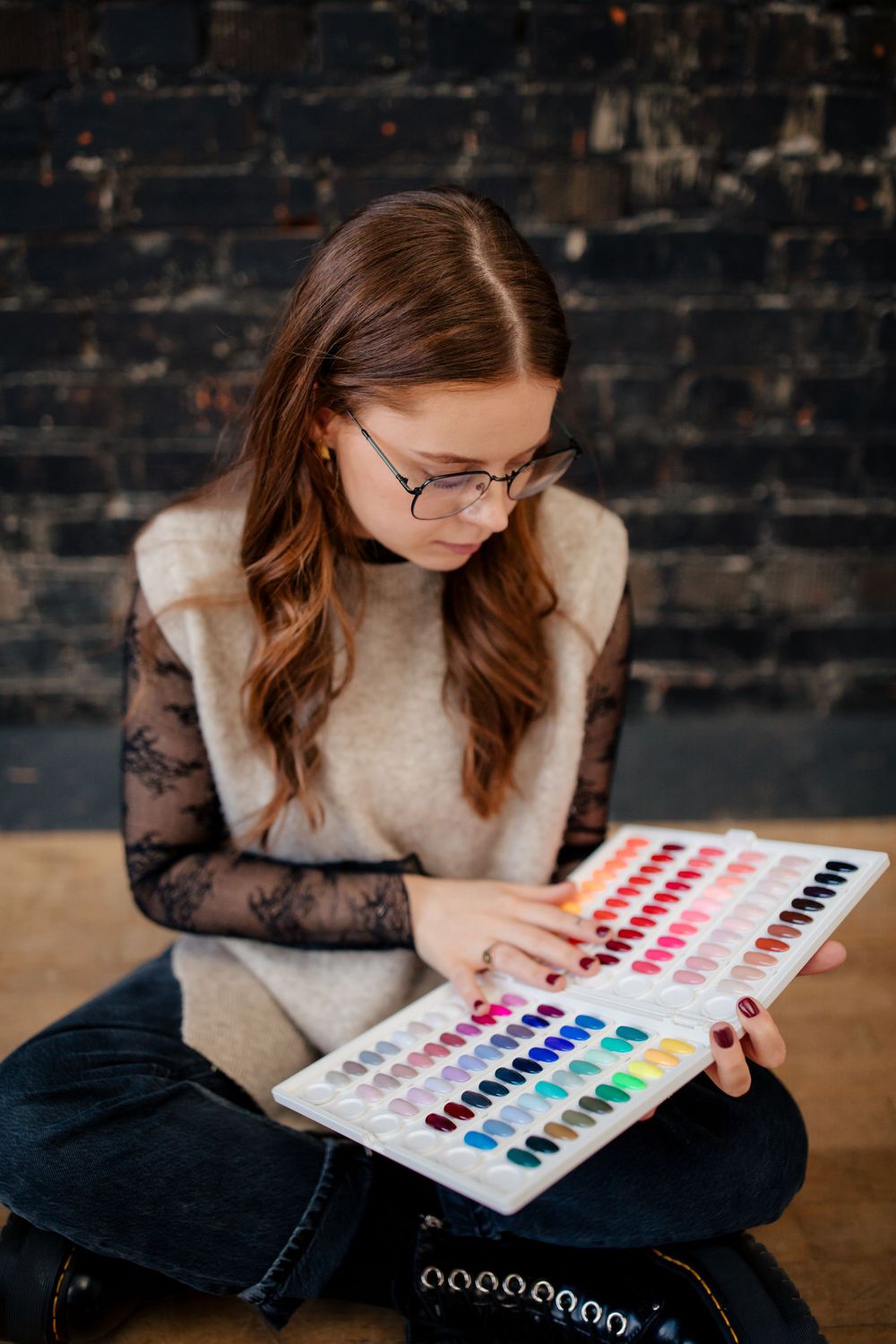 Creative artist in lace sleeves and glasses studies a colorful watercolor paint palette while sitting on wooden floor.