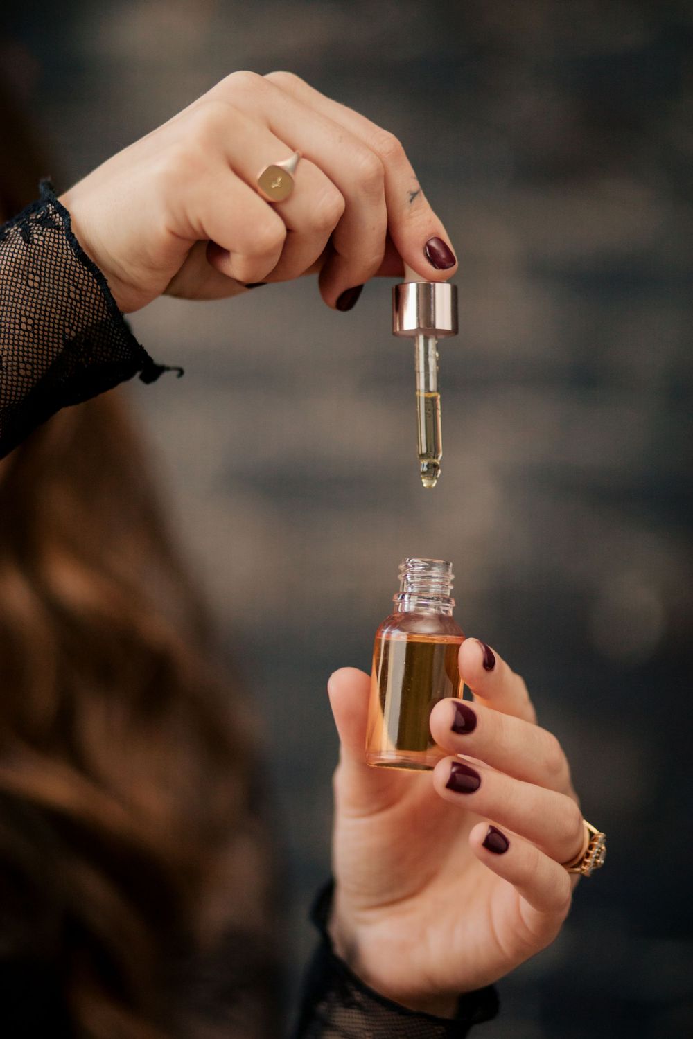A hand with dark nail polish holds a dropper of essential oil over a small glass bottle against a blurred background.