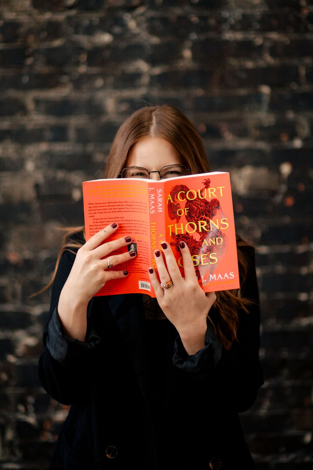 Someone in black clothing holds up an orange book titled 'A Court of Thorns' while reading behind it against a dark wall.