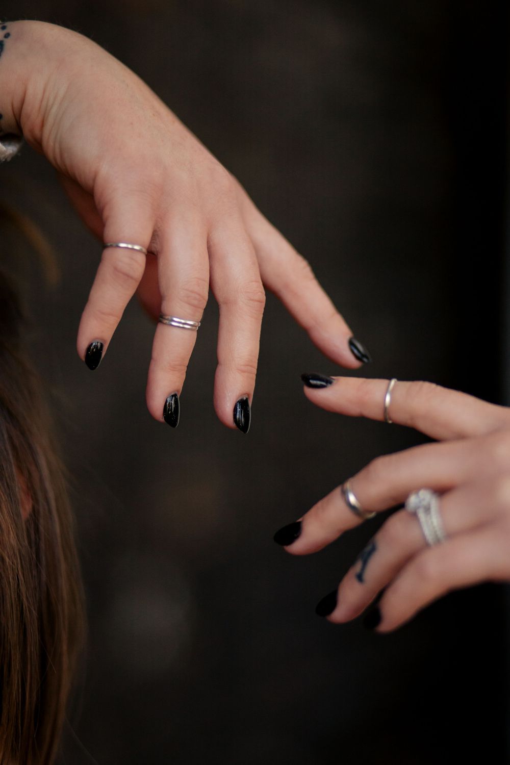 Close-up shot of hands with black nail polish wearing multiple delicate silver rings against dark background.