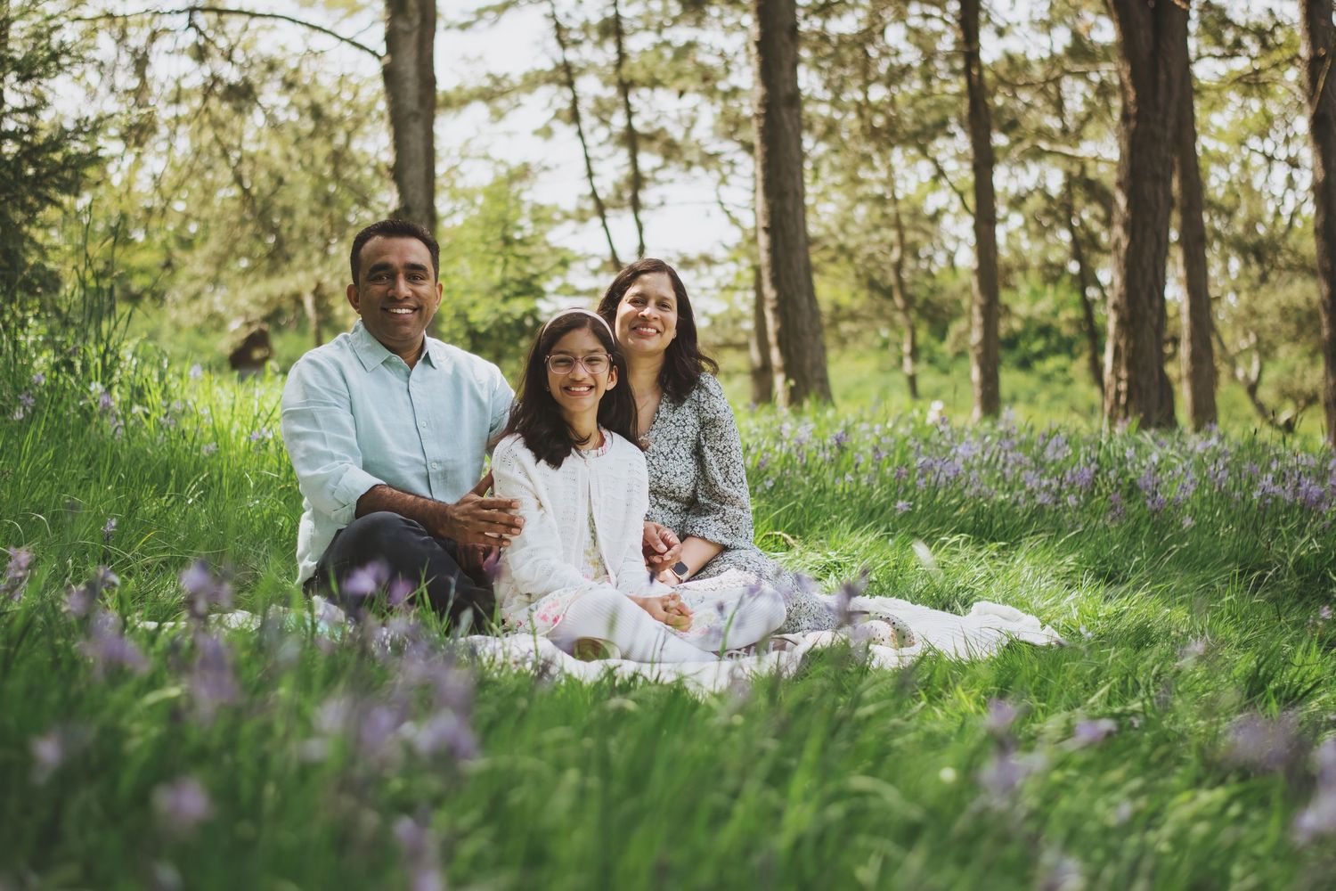 A relaxed family moment captured during a picnic in a sunlit forest meadow with blooming purple wildflowers.
