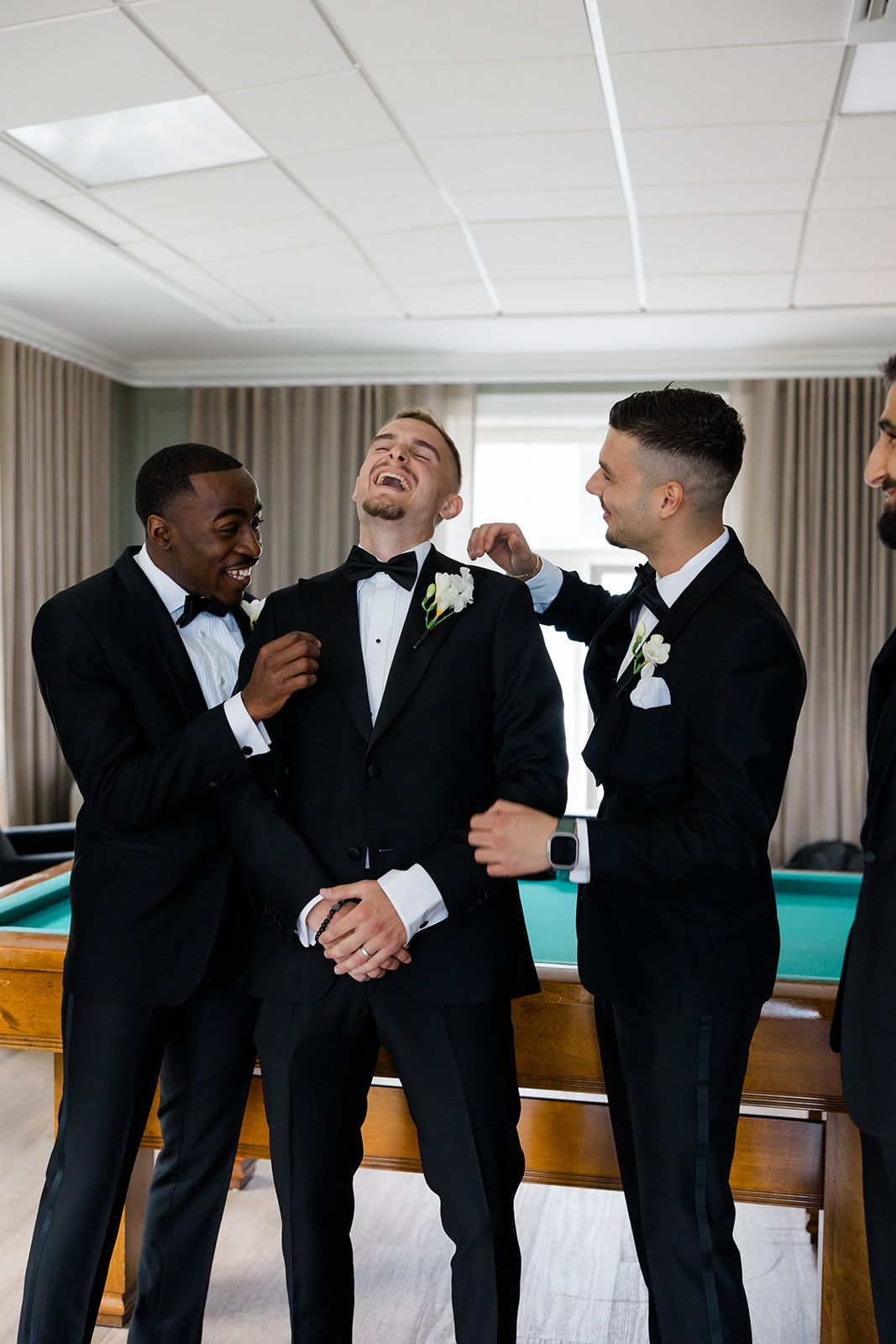 Groomsmen in black tuxedos share candid moments and laughter while getting ready before a wedding ceremony.