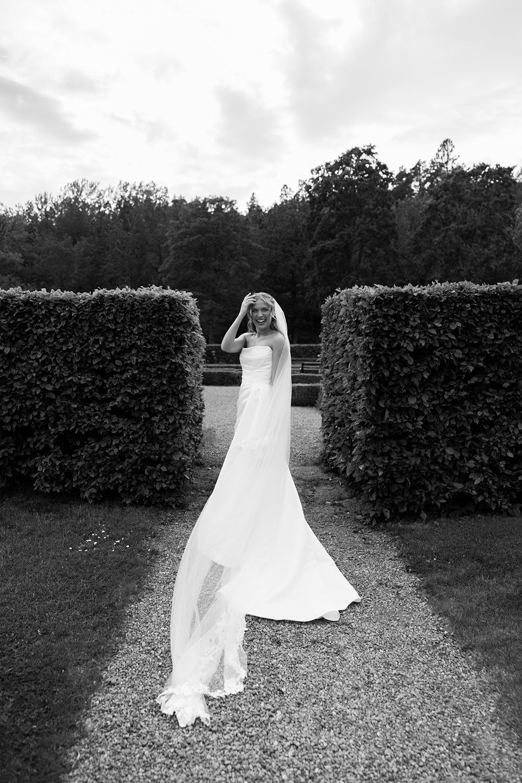A bride in a flowing white wedding dress stands between manicured hedges in a garden setting.
