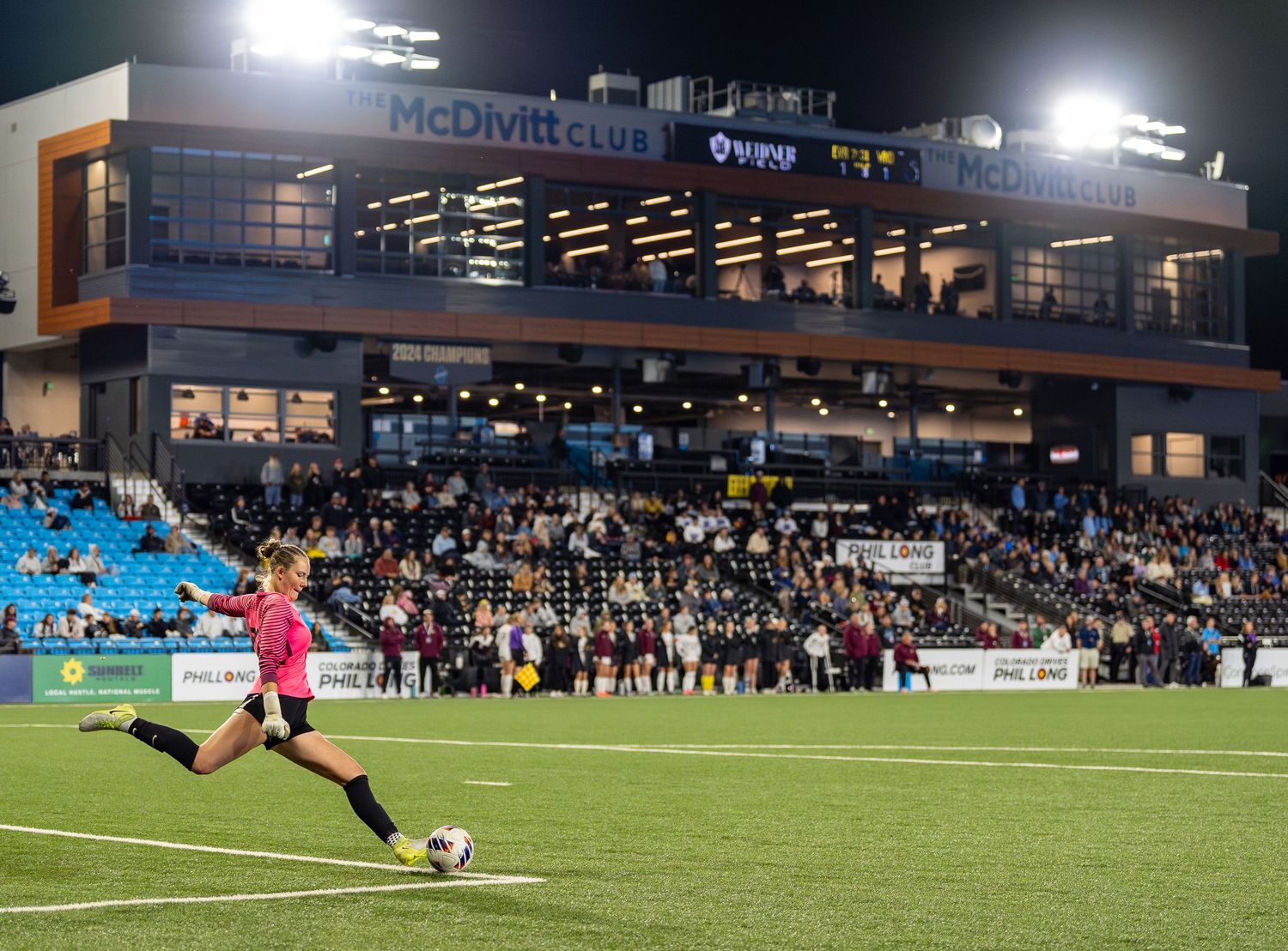 Soccer player in pink jersey takes a kick during a night game at an illuminated stadium with spectators in the stands.