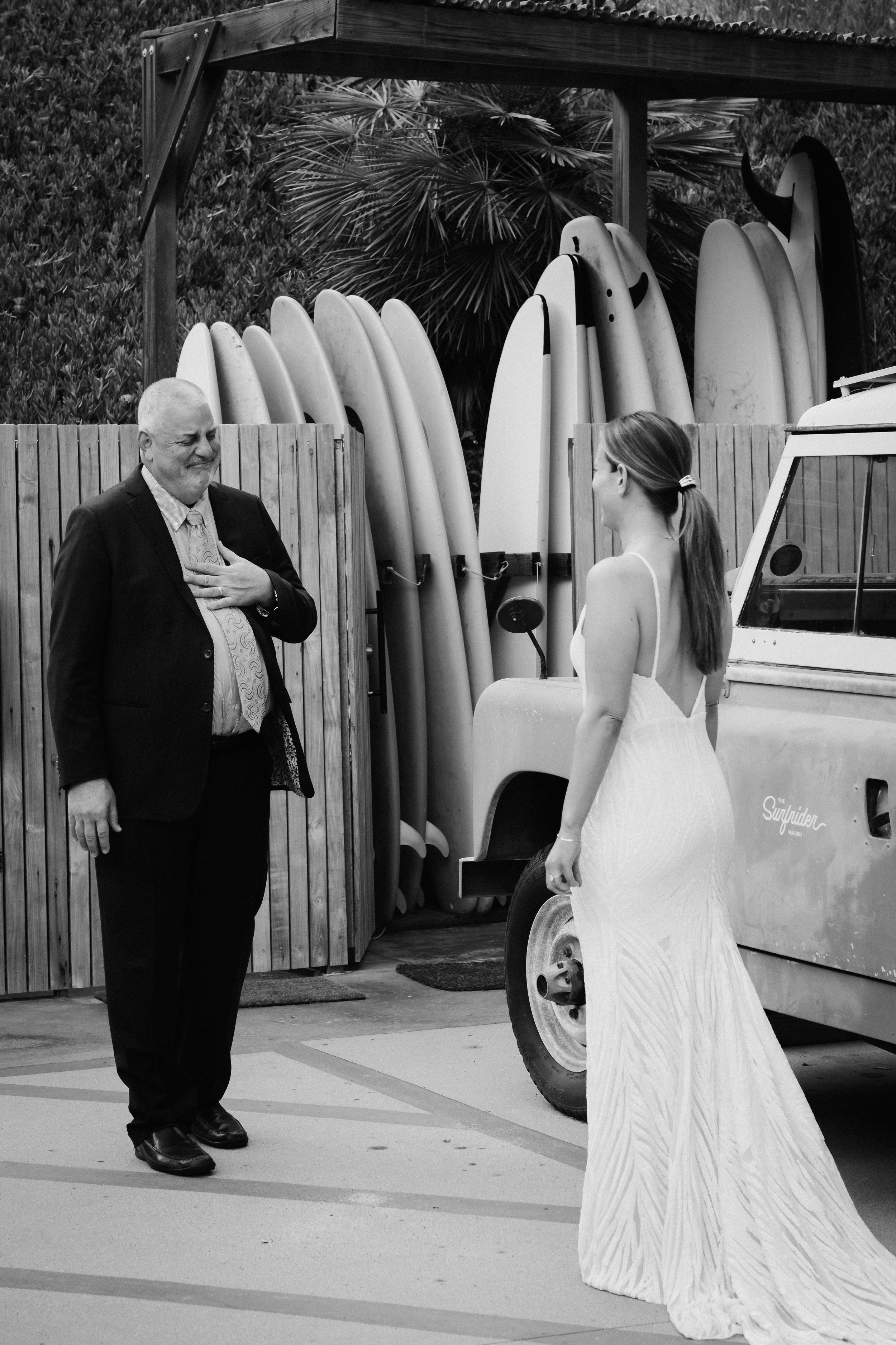 A wedding couple poses near a row of surfboards lined up against a wooden fence.