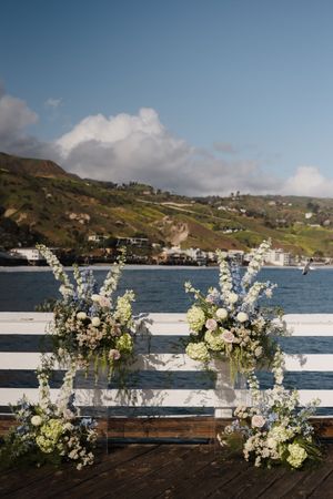 White floral arrangements line a railing with coastal mountains and blue water in the background.