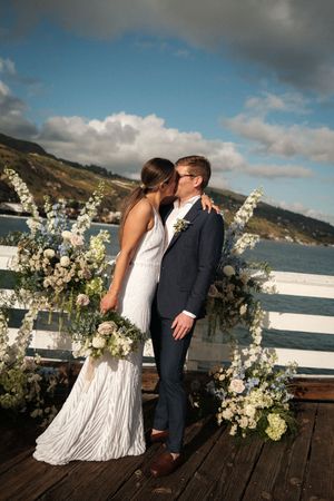 Romantic wedding couple sharing a kiss on wooden deck with white floral arrangements and ocean view backdrop.