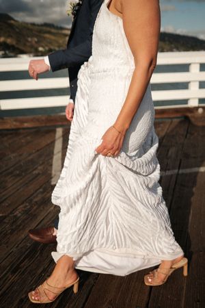 Close-up view of a white wedding dress and tan heels on a wooden deck overlooking the ocean.