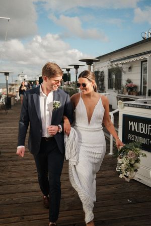 A newlywed couple walks arm in arm along a beachfront boardwalk outside Malibu restaurant on a sunny day.