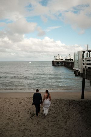 A couple walks hand in hand along a sandy beach towards a wooden pier on an overcast day.