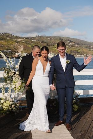 A couple celebrates joyfully on an oceanfront deck with floral decorations and scenic coastal views.