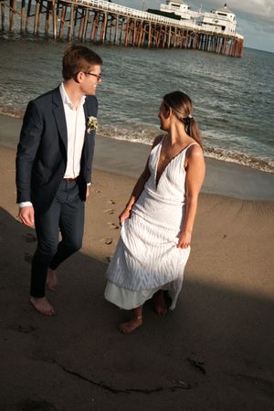 A couple walking barefoot on a beach with a pier in the background during golden hour.